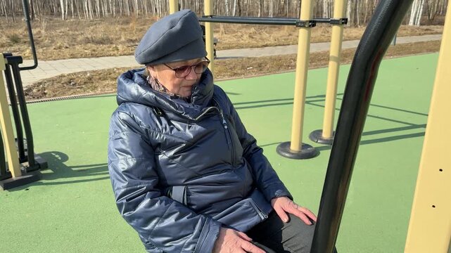 Woman exercising outdoors spring stadium wearing glasses beanie coat using leg exerciser on green rubber ground steady alternating
