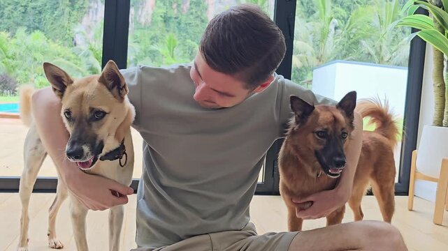 Man sitting on floor indoors holding two dogs and interacting with pets near large window with tropical garden and pool view home lifestyle with multiple animals companionship and care