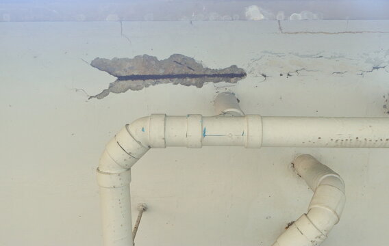 Selective focus of a significant area of concrete spalling on white ceiling, exposing a rusted rebar and aggregate within. The damage is situated directly above a network of white PVC water pipes.