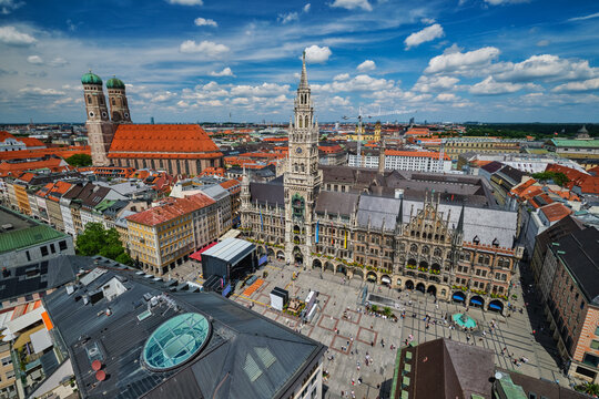Wide high-angle view of the bustling Marienplatz square, the New Town Hall, and Frauenkirche under a blue sky in Munich, Bavaria, Germany.