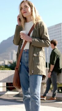 Vertical video: Glancing at smartphone, blonde woman answering call on rooftop, holding coffee