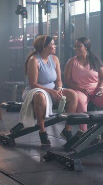 Left woman placing green bottle on bench, both laughing and gesturing while talking workout tips