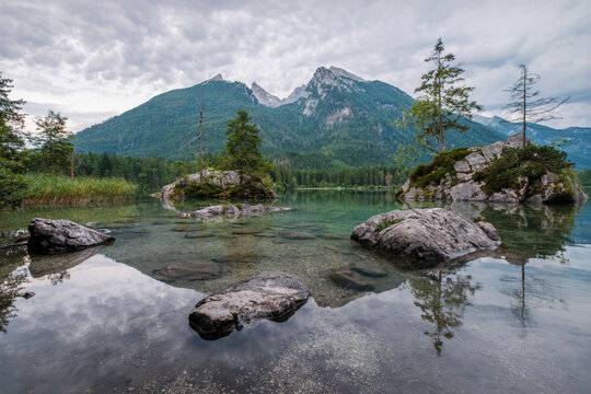 Vertical perspective of the calm waters of Hintersee lake showing rocks under the surface and the Hoher Goll mountain in Germany.