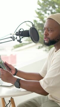 Vertical video: Moving microphone causing host in beige beanie reading tablet at window, recording