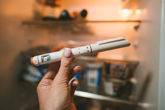 Woman hand holding GLP-1 pen in front of fridge. Semaglutide, Ozempic, Tirzepatide or insulin drug against fat. Modern weight loss treatment, diabetes therapy and obesity management