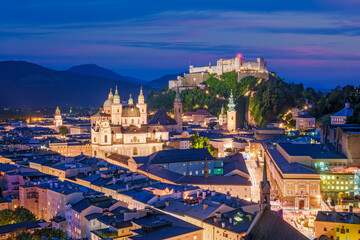 Obraz premium Close-up aerial view of the lit-up Hohensalzburg Fortress and the Salzburg Cathedral during the blue hour in the historic city center of Austria.