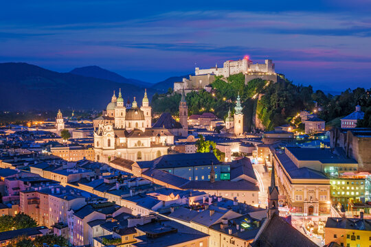 Close-up aerial view of the lit-up Hohensalzburg Fortress and the Salzburg Cathedral during the blue hour in the historic city center of Austria.