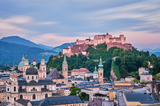 Long exposure aerial view of the illuminated Hohensalzburg Fortress and Salzburg Cathedral under a soft twilight sky at the end of the day.