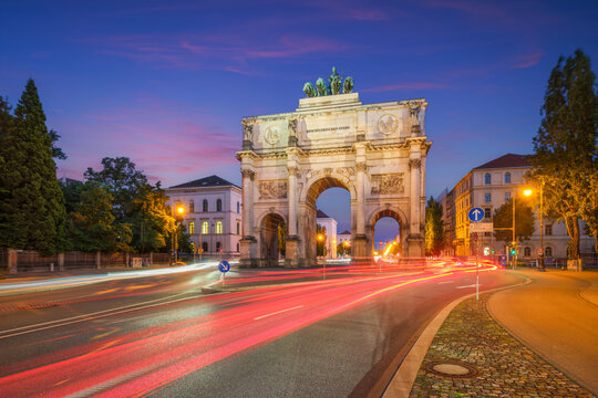 Long exposure of the illuminated Siegestor in Munich, Bavaria, Germany, featuring colorful red and white traffic light trails at blue hour.