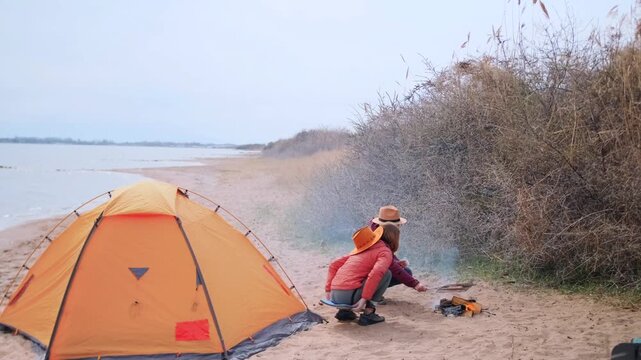 women roasting sausages over campfire crouching near tent at lakeside beach campsite, cooking food during outdoor trip, enjoying camping routine, travel lifestyle and leisure time by water