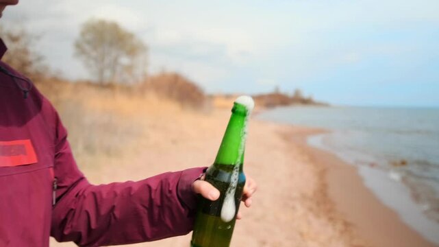 Person opening beer bottle on lakeside beach, relaxing during outdoor travel, camping vacation, casual lifestyle moment, drink in nature, vacation atmosphere, leisure time near water