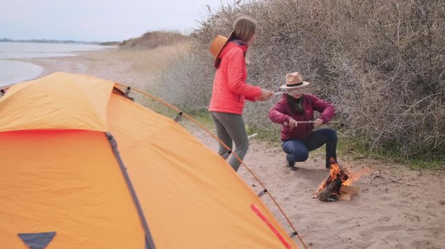 woman tending campfire crouching near tent at lakeside beach campsite, blowing on fire to ignite wood while friend stands nearby, outdoor camping routine and travel lifestyle by water