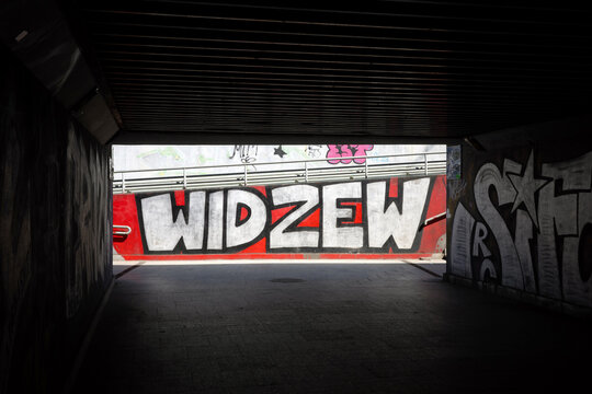 LODZ, POLAND - APRIL 18, 2026: A street level photograph captures a prominent white graffiti mural reading "WIDZEW" on a red wall within an urban underpass, taken with direct sunlight.