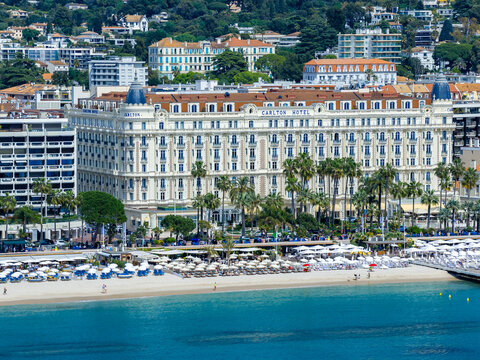 Aerial Cannes, France, shows Carlton Hotel facing turquoise water and sandy beach, palm lined promenade, umbrellas, and loungers in bright daylight during festival season.