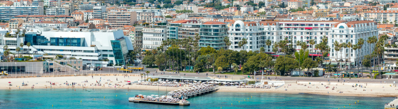 Aerial view of Cannes, France, shows Croisette beach, a sunbed pier, the Palais des Festivals on the left, and the Carlton Cannes to the right under bright daylight.