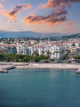 Aerial view of the Bay of Cannes and La Croisette in France shows the Carlton hotel, sandy beach, marina, palm trees, and city hills under partly cloudy daylight skies.