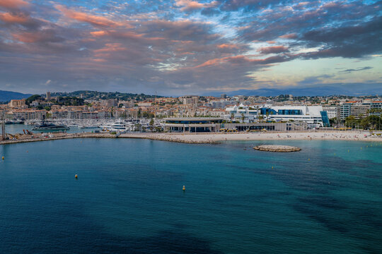Aerial view of Cannes, France shows the Bay of Cannes, sandy beach, Vieux Port yachts, yellow buoys, and the Palais des Festivals, with terraced city and hills in sun.