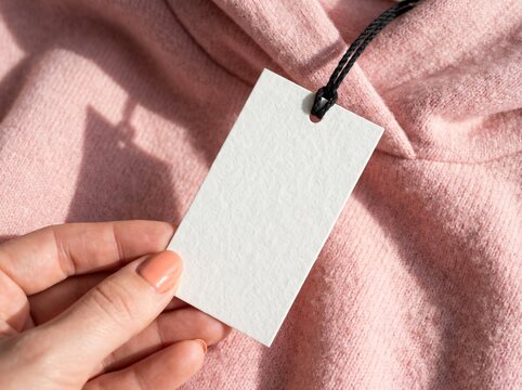 Hand holding blank white tag attached to a pink hoodie mockup for clothing label design fashion product branding soft texture close-up