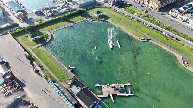 4K aerial drone footage of Hove Lagoon featuring windsurfing, cable tow wakeboarding and sailing. Vibrant coastal watersports action on the sunny Sussex seafront.
