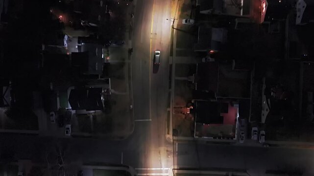 Aerial, top down view tracking a car driving through a suburban Long Island, NY neighborhood at night. Capturing streetlights in a surveillance-style shot as the car follows a bend in the road.