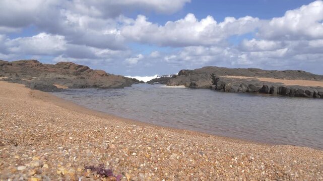 A serene coastal scene unfolds with clear water rippling over a textured pebble shore, and waves crash against dark rocks, set against a backdrop of dramatic clouds at Ramsgate, South Coast.