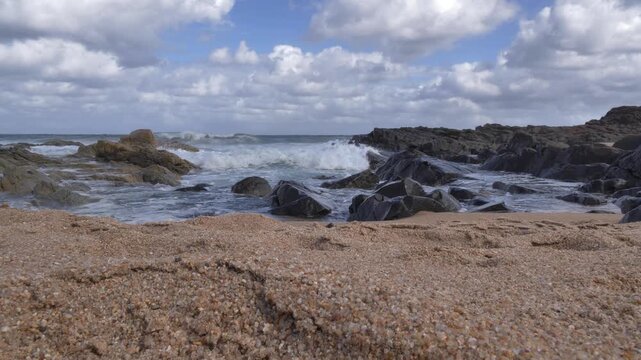 The scene moves from light to shade as ocean waves surge and break against dark, rugged rocks and a textured sandy shore under a dramatic sky filled with clouds at Ramsgate, South Coast, KwaZulu-Natal