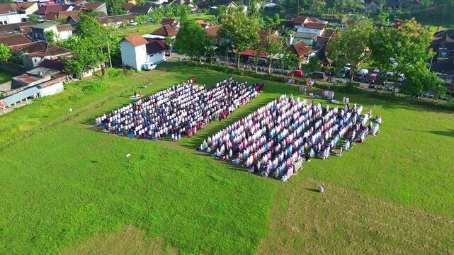 Large group of Muslims in sujood during Eid al-Fitr prayer, captured from above on a green open field.