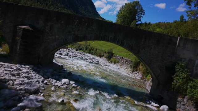 Aerial shot of a stone arch bridge spanning a fast mountain river in the Bregaglia valley.