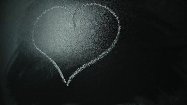 A hand draws a heart shape on a chalkboard with white chalk.