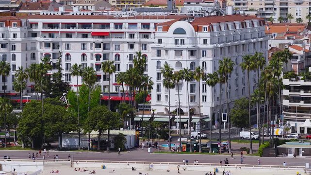 Aerial view of La Croisette in Cannes with InterContinental Carlton hotel, palm promenade, sandy beach, umbrellas, sunbathers, pedestrians, and gentle traffic motion.