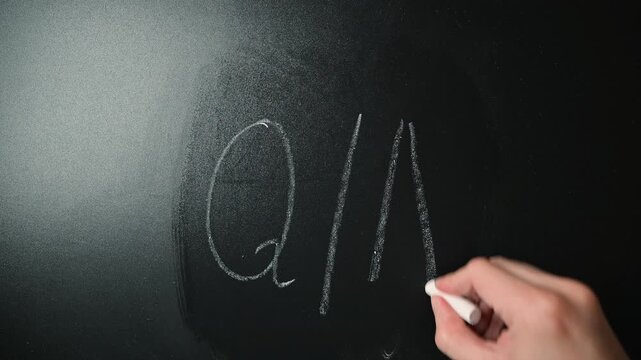 A hand writes letters and symbols on a blackboard using chalk in a classroom.
