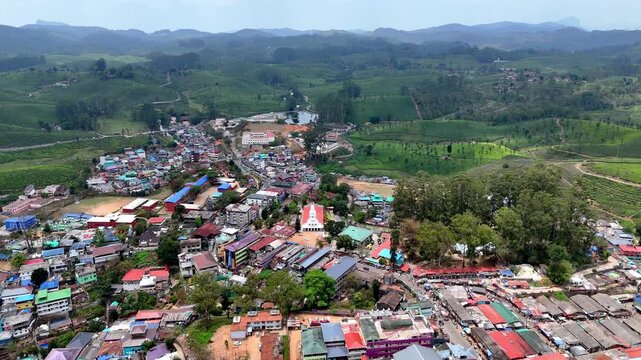 Cinematic aerial view of a vibrant hill town with colorful rooftops, a prominent Church structure at the center, and dense clusters of buildings, surrounded by expansive green plantations and rolling