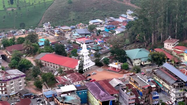 Cinematic aerial view of a hillside town featuring colorful rooftops, clustered buildings, and a prominent white Church at the center, surrounded by trees and rolling terrain with a calm, small-town.