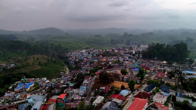 Cinematic aerial view of Valparai showcasing a dense hill town with colorful rooftops, winding streets, and open grounds, surrounded by rolling tea plantations and misty hills.