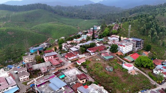 Drone shot capturing lush tea estates in Valparai with a cluster of vibrant houses nestled at the edge of the plantations, surrounded by forested hills and curving roads.