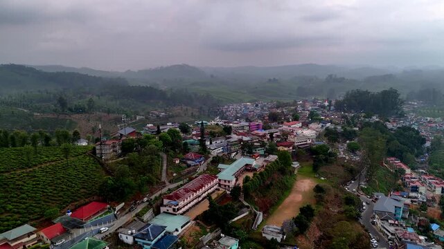 Drone shot capturing Valparai with compact housing clusters, busy roads, green hills, and layered mountain ranges under a soft hazy sky.