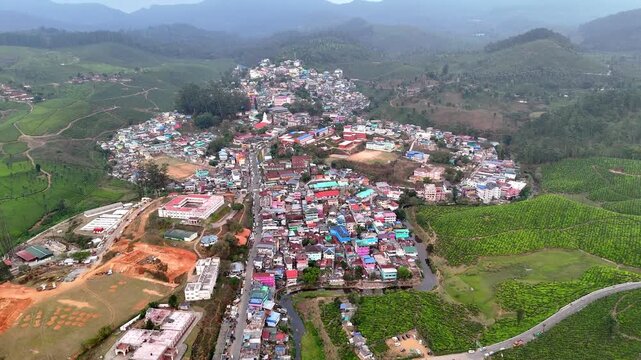 Wide cinematic aerial of Valparai highlighting textured tea gardens, a serpentine trail, and a compact settlement blending into dense forest cover and rolling hills. curving hillside roads