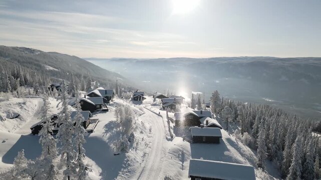 Aerial winter view of a Scandinavian cabin village in Danebu, Valdres, Norway, covered in falling snow, with bright, morning sunlight flaring over the Nordic retreat