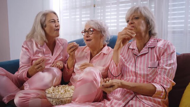 Beautiful senior women having funny pajama party at home - Group of cheerful elderly female friends eating pop-corn while watching series on tv, concepts about old people, lifestyle, quality of life	