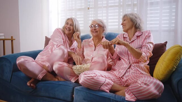 Beautiful senior women having funny pajama party at home - Group of cheerful elderly female friends eating pop-corn while watching series on tv, concepts about old people, lifestyle, quality of life	