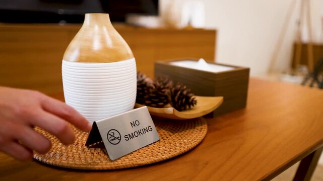 A person placing a no smoking sign on a table next to a decorative vase, wooden box, and pine cones.