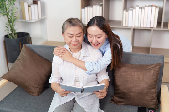Asian mother and daughter hugging, reading book together in home living room