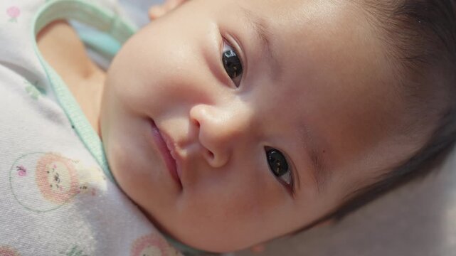 Soft-focus close-up of a calm baby's face. Big, dark eyes look gently toward the camera. Tiny nose and soft lips add to the serene expression. Baby rests on a light-colored, patterned blanket