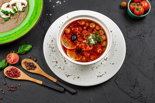 Traditional russian solyanka soup with sausages olives and lemon. Top view of meat soup with olives lemon and cream in white bowl on dark stone background.