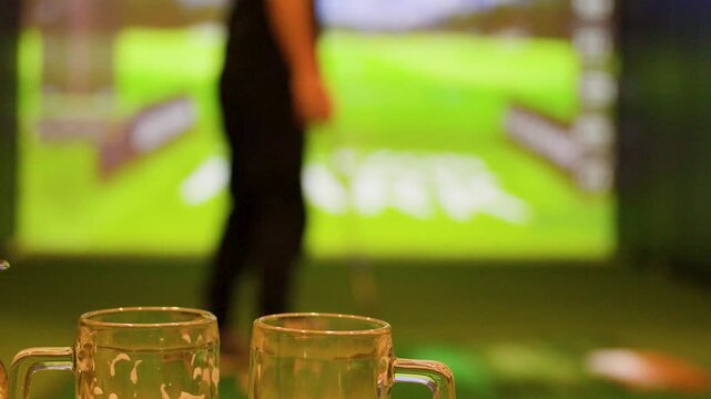 Caucasian Man Swinging Golf Club in Indoor Simulator Near Beer Mugs