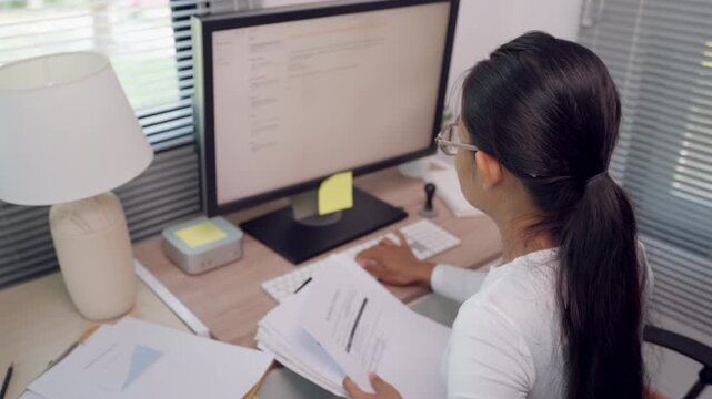 Female office worker reviewing printed documents at desk beside computer monitor. Business administration, paperwork management, document checking, and professional office workflow concept.