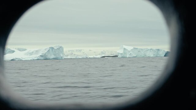 Row of flat tabular Antarctic icebergs on grey polar ocean viewed through oval ship porthole illuminator window