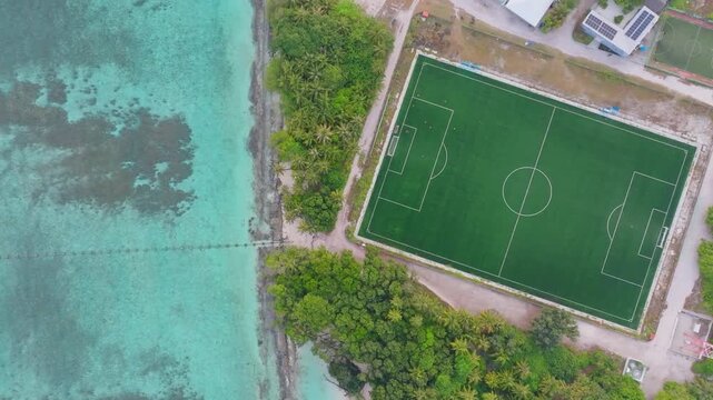Top down aerial drone footage over the eastern side of Vashafaru Island featuring the local football field and homes of the island community in northern Maldives.
