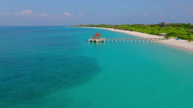 Scenic aerial drone flyover along the white sand beach of Vashafaru Island featuring turquoise water, a long jetty and a covered pavilion with multiple water access points.