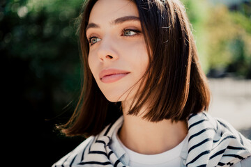 Fototapeta premium Young woman portrait outdoors looking thoughtful with short brown bob, green eyes and striped jacket in soft sunlight